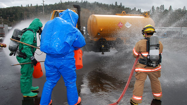 Two people wearing hazmat suits with a firefighter cleaning up a flammable chemical spill