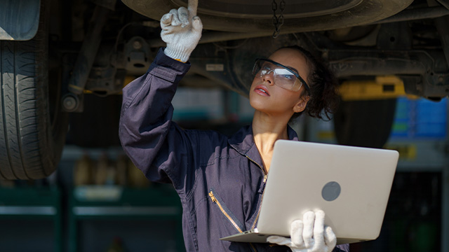 Female mechanic lone worker underneath car with laptop and personal protective equipment on