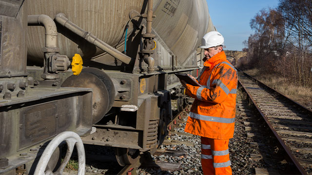 A railway maintenance Inspector wearing high visibility clothing and protective safety work wear examining a tanker wagon at the track side